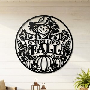 May include: A circular black metal wall art piece featuring a scarecrow wearing a hat with a sunflower, surrounded by wheat stalks and pumpkins. The center text reads HELLO FALL in bold lettering. The design is a cutout silhouette style.