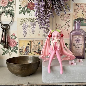 May include: A pink anime figurine with long pigtails and cherry-shaped hair accessories sits on a concrete surface. A vintage purple bottle with "Blossom Tonic" text is behind the figurine. The background includes floral art prints and a bronze bowl.