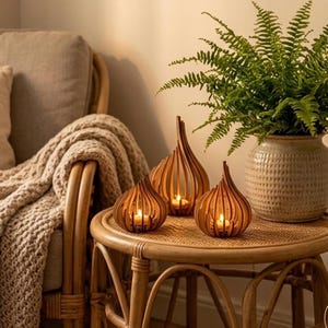 Peut inclure: Trois bougeoirs en bois en forme de goutte d'eau, chacun contenant une bougie allumée. Les bougeoirs sont posés sur une table ronde en osier, à côté d'une fougère dans un vase texturé. Un fauteuil et une couverture en osier sont en arrière-plan.