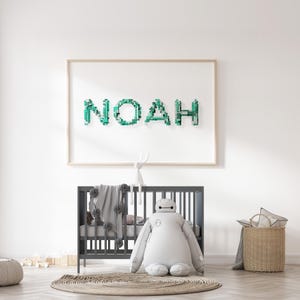 Könnte beinhalten: Ein gerahmtes Kunstwerk mit dem Namen "NOAH" in türkisfarbenen Bausteinen hängt über einem grauen Kinderbett. Ein großes Plüschroboterspielzeug und ein Weidenkorb befinden sich im Vordergrund. Eine graue Decke mit Bommeln ist über das Kinderbett drapiert.
