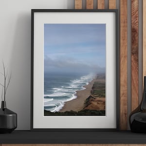 Peut inclure: Photographie encadrée d'un paysage côtier. L'image présente une longue plage de sable, des vagues océaniques et un ciel nuageux. La photographie est dans un cadre noir et est exposée sur une surface en bois.
