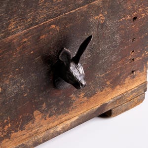Puede incluir: Un cofre de madera rústico con un tirador en forma de cabeza de ciervo negro. El cofre tiene un aspecto envejecido y desgastado con vetas de madera visibles y un acabado marrón oscuro. La cabeza de ciervo es una escultura tridimensional detallada.