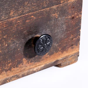 May include: A rectangular wooden box with a weathered, dark brown finish showing visible wood grain and texture. A single circular black metal knob is attached to the front, featuring an embossed floral pattern with symmetrical curved lines.