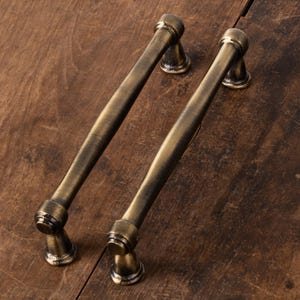 May include: Two matching cabinet pulls with a dark antique brass finish. Each handle features a long, cylindrical bar with a smooth, slightly tapered centre and rounded ends, supported by two flared, circular mounting posts at each side.