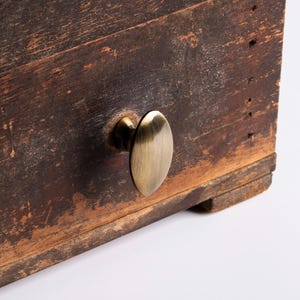May include: Close-up of a distressed wooden drawer featuring an oval brass knob. The wood shows signs of age and wear, with visible grain and imperfections. The brass knob is centrally positioned, offering both function and decoration.