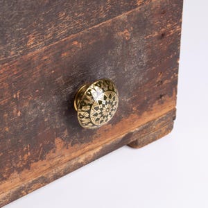 Könnte beinhalten: Eine dunkelbraune, verwitterte Holzschubladenfront mit einem runden Metallknopf. Der Knopf hat eine polierte goldfarbene Oberfläche mit einem schwarzen, komplizierten, geometrischen und blumenartigen Muster, das in die Oberfläche eingraviert ist.