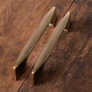 May include: Two antique brass cabinet pulls with a sleek, modern design. The pulls are rectangular with a slightly curved handle, and are set against a rustic, wooden surface. These hardware pieces are ideal for updating kitchen or bathroom cabinets.