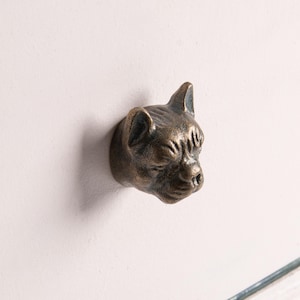 May include: A bronze-toned metal cabinet knob shaped like the head of a French Bulldog. The knob features detailed facial features including pointed ears, a wrinkled forehead, and a short snout, providing a three-dimensional sculptural appearance.