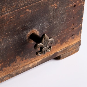 Pode incluir: Close-up de uma caixa de madeira desgastada com uma pega decorativa de metal. A pega tem a forma de uma flor-de-lis e está presa à caixa com uma haste preta. A madeira tem um aspeto envelhecido e desgastado.