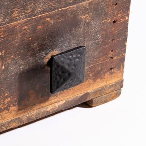 Pode incluir: Close-up de uma seção de uma caixa de madeira rústica com uma alça de metal preta, quadrada e texturizada. A madeira tem uma aparência desgastada com grãos visíveis e variações de cor, variando de marrom claro a marrom escuro.