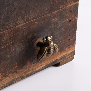 May include: Close-up of a rustic wooden box with a bronze bee-shaped handle. The box has a distressed, aged appearance with visible wood grain and a dark brown color. The bee handle adds a decorative touch.