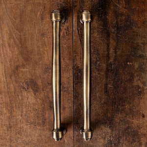 Puede incluir: Dos tiradores de armario de latón antiguo con un diseño cilíndrico vertical. Los tiradores están montados sobre una superficie de madera marrón oscura y envejecida, creando una estética vintage. El herraje tiene un aspecto clásico y elegante.