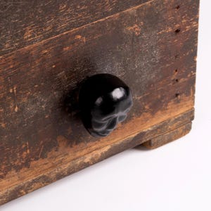 May include: A dark brown wooden drawer front with a weathered, distressed finish and a single, glossy black cabinet knob shaped like a human skull. The wood grain is visible, showing signs of age and wear across the rectangular surface.