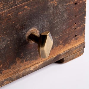 May include: Close-up of a weathered wooden surface with a brass-coloured, geometric cabinet knob. The wood has a distressed appearance with visible grain and dark brown tones. The knob is diamond-shaped.
