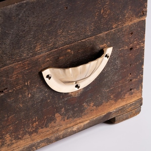 Può includere: Primo piano di una scatola di legno invecchiato con una maniglia in ottone. La scatola è realizzata in legno marrone scuro, usurato, con venature e fori di chiodi visibili. La maniglia in ottone curva è fissata con tre viti, aggiungendo un tocco di eleganza.