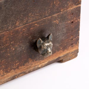 May include: A dark brown, weathered wooden box featuring a single metallic drawer pull shaped like the head of a French Bulldog. The wood surface shows signs of age, including visible grain, scratches, and a worn, distressed texture throughout.
