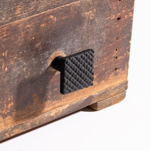 Puede incluir: Primer plano de una caja de madera envejecida con un tirador cuadrado negro. El tirador tiene una superficie texturizada con un patrón de diamantes. La madera muestra signos de edad con vetas visibles y un acabado desgastado, lo que sugiere una estética vintage o rústica.