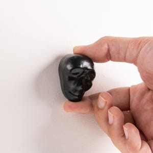 May include: A small, smooth, black, skull-shaped object with a glossy finish. The skull features defined eye sockets, a nasal cavity, and a subtle jawline, presented as a singular, compact decorative piece with a rounded, polished surface texture.