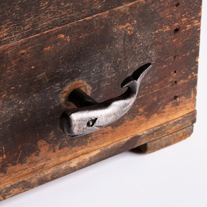 May include: A rustic wooden box with a whale-shaped handle. The handle is made of metal and is attached to the box with a metal rod. The box is made of weathered wood with visible grain and knots.