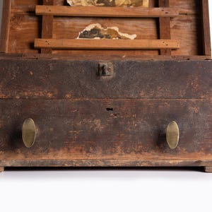 May include: A weathered wooden chest with a dark, distressed finish, featuring two oval brass knobs on the front drawer, a central metal latch, and an open lid revealing internal wooden slats and remnants of aged paper documents inside.