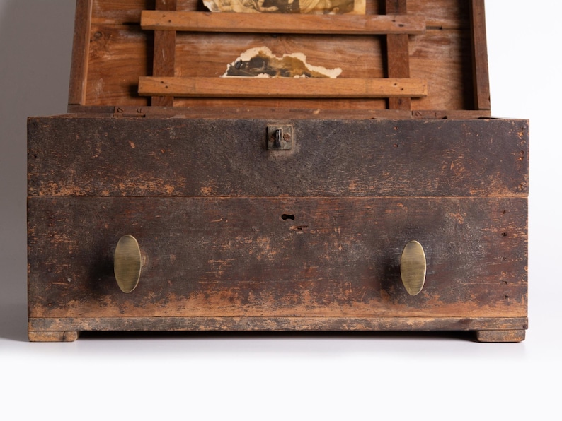 May include: A weathered wooden chest with a dark, distressed finish, featuring two oval brass knobs on the front drawer, a central metal latch, and an open lid revealing internal wooden slats and remnants of aged paper documents inside.