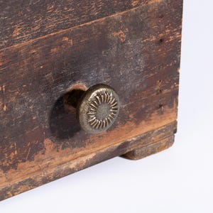 May include: A dark brown wooden drawer front with a weathered, distressed finish and visible wood grain. A round, antique brass-toned metal knob is attached to the center, featuring a textured, sunburst-like radial pattern on its face.