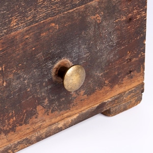 May include: A dark brown wooden drawer front featuring a weathered, distressed finish with visible wood grain and texture. A single round, metallic brass-toned knob is centered on the front panel, providing a contrast to the aged wood surface.
