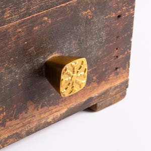Puede incluir: Una superficie de madera oscura y desgastada presenta un pomo cuadrado de latón. El pomo tiene un acabado dorado pulido y reflectante, con un patrón texturizado en relieve en su cara frontal que se asemeja a un copo de nieve estilizado.