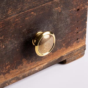 Puede incluir: Primer plano de un tirador de armario dorado con un diseño circular texturizado. El tirador está montado sobre una superficie de madera marrón oscura y envejecida, mostrando el contraste entre lo nuevo y lo antiguo.