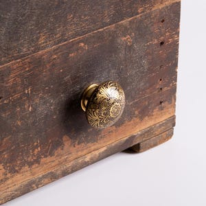 Pode incluir: Close-up de um baú de madeira desgastada com um puxador floral decorativo dourado e preto. O baú tem uma aparência rústica e envelhecida com grão de madeira e imperfeições visíveis, sugerindo uma peça vintage ou antiga.