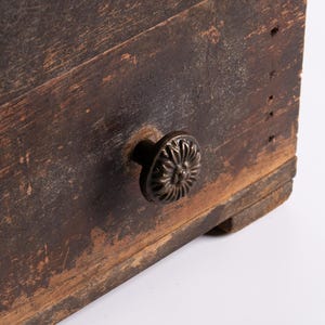 Op de afbeelding: Close-up van een verweerde houten lade met een decoratieve, donkere metalen knop. Het hout heeft een verweerde uitstraling met zichtbare nerven en een rijke, bruine kleur, wat wijst op een antieke of rustieke stijl.