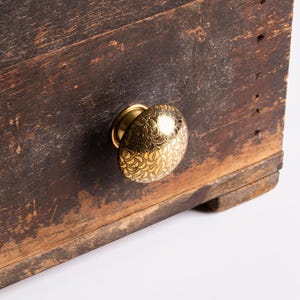 Puede incluir: Primer plano de un cajón de madera con un pomo dorado decorativo. El pomo tiene un patrón intrincado y está montado sobre una superficie de madera marrón oscura y desgastada. La madera muestra signos de edad y desgaste.