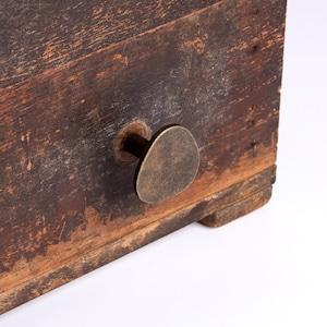 May include: A dark brown wooden drawer featuring a weathered, distressed finish with visible wood grain and texture. A single, asymmetrical, antique brass-toned metal knob is attached to the front, providing a contrast to the aged wood surface.
