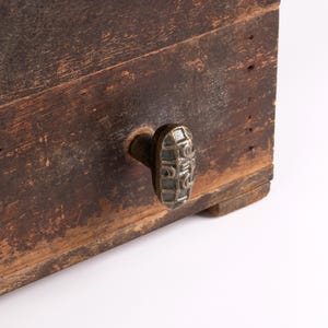 May include: A dark brown wooden box featuring a weathered, distressed finish with visible wood grain. The box is equipped with a single oval-shaped metal knob that has a textured, grid-like pattern on its front surface and a dark metallic finish.