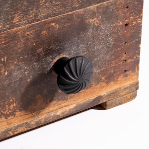 Puede incluir: Primer plano de una caja de madera envejecida con un pomo negro en espiral. La madera tiene un aspecto desgastado con vetas visibles y variaciones de color. El pomo está centrado en la parte frontal de la caja.