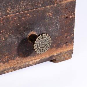 Könnte beinhalten: Eine verwitterte, dunkelbraune Holzschubladenfront mit einem runden Messingknopf, der ein detailliertes, strahlenförmiges Blumen- oder Zahnradmuster aufweist. Die Holzoberfläche zeigt deutliche Altersspuren mit sichtbarer Maserung und Textur.