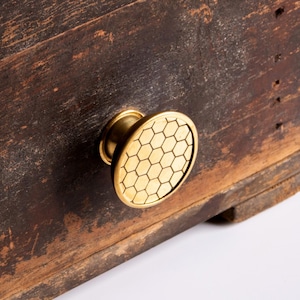 May include: A round brass cabinet knob featuring a geometric honeycomb pattern on its face. The knob is attached to a dark, weathered wooden surface with visible grain and signs of age, showcasing a contrast between the metallic finish and wood.