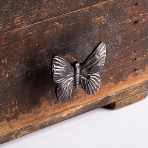 Peut inclure: Poignée de tiroir en métal argenté en forme de papillon, fixée sur une surface en bois brun foncé vieilli. Le papillon présente des motifs détaillés sur les ailes. Le bois montre des veines et des trous de clous visibles, pour un aspect rustique.