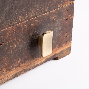 May include: A rectangular, dark brown, weathered wooden box featuring a single, vertically oriented, brushed brass cabinet knob attached to the front panel. The wood surface shows visible signs of age, including scratches and worn texture.
