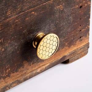 May include: Close-up of a wooden cabinet with a gold-colored, honeycomb-patterned knob. The cabinet has a distressed, dark brown finish, showing signs of wear and age. The knob is circular and adds a touch of elegance to the rustic design.