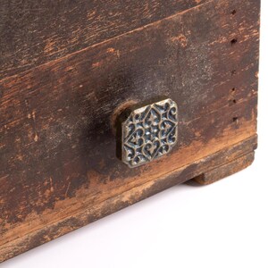Könnte beinhalten: Eine dunkelbraune, verwitterte Holzschubladenfront mit einem quadratischen Metallknopf. Der Knopf hat eine silberfarbene Oberfläche mit einem komplexen, erhabenen und symmetrischen Filigranmuster aus geschwungenen Linien und herzartigen Formen.