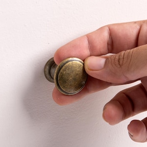 May include: A round cabinet knob with an antique brass finish, featuring a smooth, slightly domed surface and a raised circular border detail. The knob has a cylindrical base that connects to the back, showing a weathered, metallic texture.