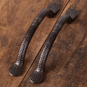 Op de afbeelding: Twee donker bronzen kastgrepen met een gehamerde textuur. De handgrepen hebben een gebogen ontwerp met een getextureerde grip en afgeronde uiteinden. De hardware is geplaatst op een rustiek houten oppervlak.