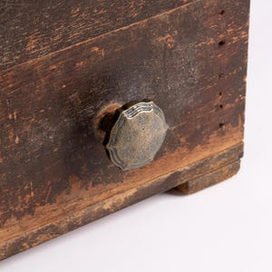Puede incluir: Un frente de cajón de madera marrón oscuro y desgastada, con un acabado envejecido que muestra la veta y textura de la madera. Un pomo metálico redondo con borde festoneado y una pátina de tono latón antiguo está centrado en el frente.