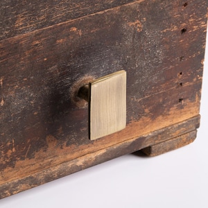 May include: Close-up of a weathered wooden box with a rectangular brass handle. The wood has a dark brown, distressed finish with visible grain and imperfections. The brass handle is mounted on the front, adding a touch of contrast.