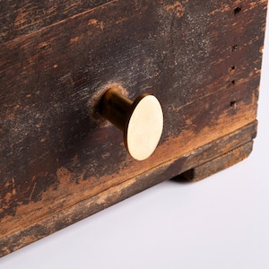 Könnte beinhalten: Nahaufnahme einer verwitterten Holzkiste mit einem Messingknauf. Das Holz hat ein dunkles, gealtertes Aussehen mit sichtbarer Maserung und Unvollkommenheiten. Der runde Messingknauf bietet einen kontrastierenden, modernen Akzent.