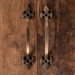 Könnte beinhalten: Zwei antike Messing-Schrankgriffe mit einem pfeilförmigen Design. Die Griffe sind auf einer verwitterten Holzoberfläche montiert und zeigen eine Vintage-Ästhetik. Die Hardware verfügt über einen langen, schlanken Griff mit aufwendigen Details.