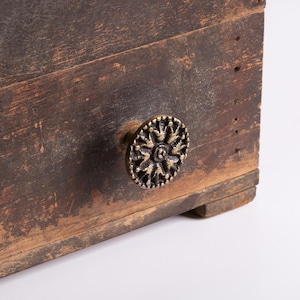 以下が含まれることがあります： アンティーク調の真鍮製ノブが付いた、風化した木製のチェストのクローズアップ。木材は、木目や釘穴が見え、古さと素朴な魅力を醸し出しています。ノブは円形で、華やかな花の模様が施されています。