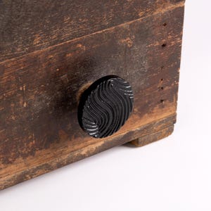May include: A dark brown, weathered wooden drawer front featuring a circular black knob. The knob has a glossy finish and a distinct, raised wavy or swirling texture across its surface. The wood shows signs of age with visible grain and wear.