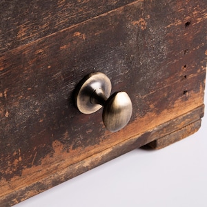 May include: Close-up of a weathered wooden box with a brass-colored knob. The wood has a distressed appearance with visible grain and variations in color. The knob is oval-shaped and mounted on a circular base.